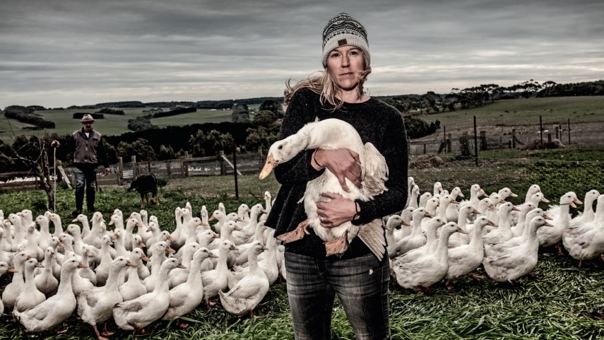 An unsmiling woman in a beanie. jumper and jeans holds a white duck as she stands with a flock of similar birds in a paddock