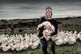 An unsmiling woman in a beanie. jumper and jeans holds a white duck as she stands with a flock of similar birds in a paddock