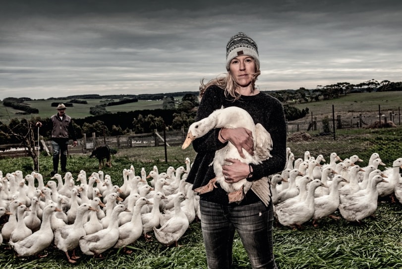 An unsmiling woman in a beanie. jumper and jeans holds a white duck as she stands with a flock of similar birds in a paddock