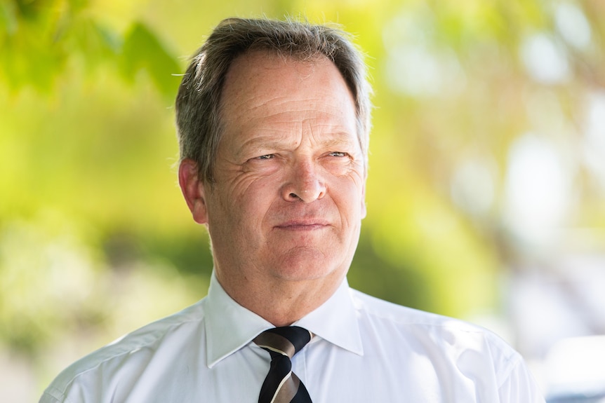 A man in a white shirt with a black and gold tie stands outside looking into the distance.