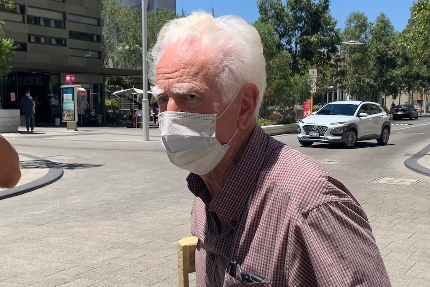 A tight head and shoulders shot of an elderly man with white hair and a face mask walking in front of a street with a car on it.