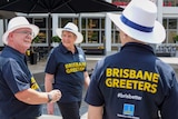 Three older people walk together wearing white hats and navy polos that read "Brisbane Greeters".