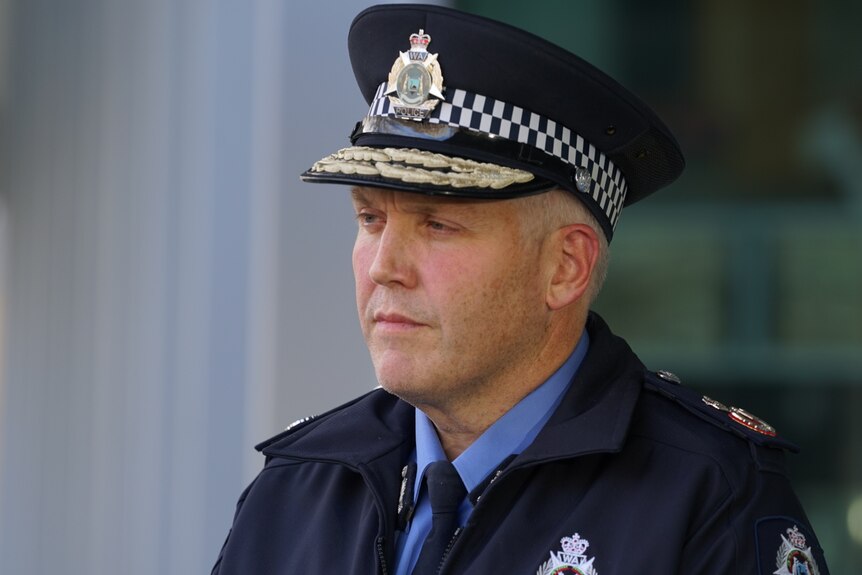 A middle-aged policeman in uniform stands outdoors, looking serious.