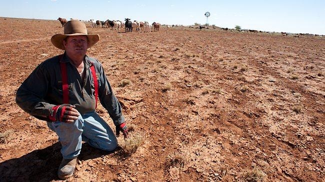 Mayor of Boulia Rick Britton squats front of frame with dry earth and windmill behind him