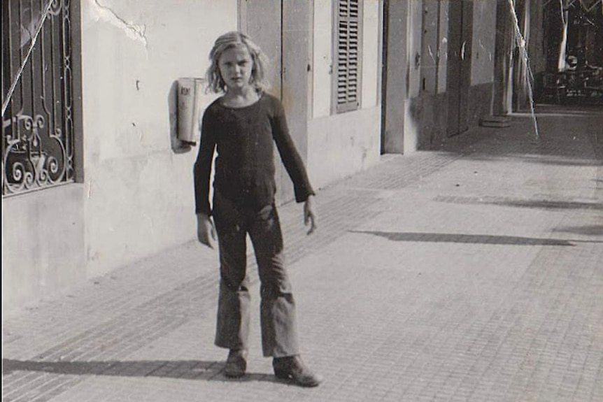 A black and white photo of a boy with long blonde hair standing on a street and looking at the camera.