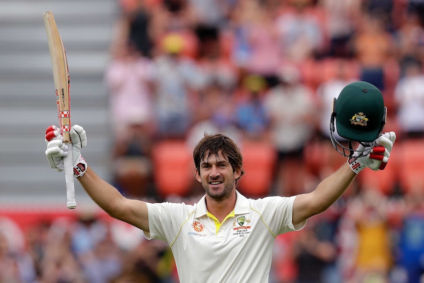 A man stands with his arms aloft, a cricket bat in his right hand, a batting helmet in his left.