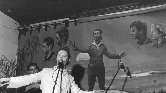 Dave Graney with outstretched arms stands at a microphone in front of a mural wearing a white suit