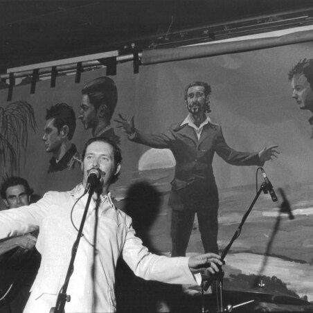 Dave Graney with outstretched arms stands at a microphone in front of a mural wearing a white suit