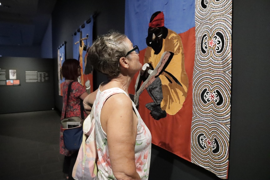 Two women look at textile banners depicting Aboriginal people. They are on the wall of a gallery