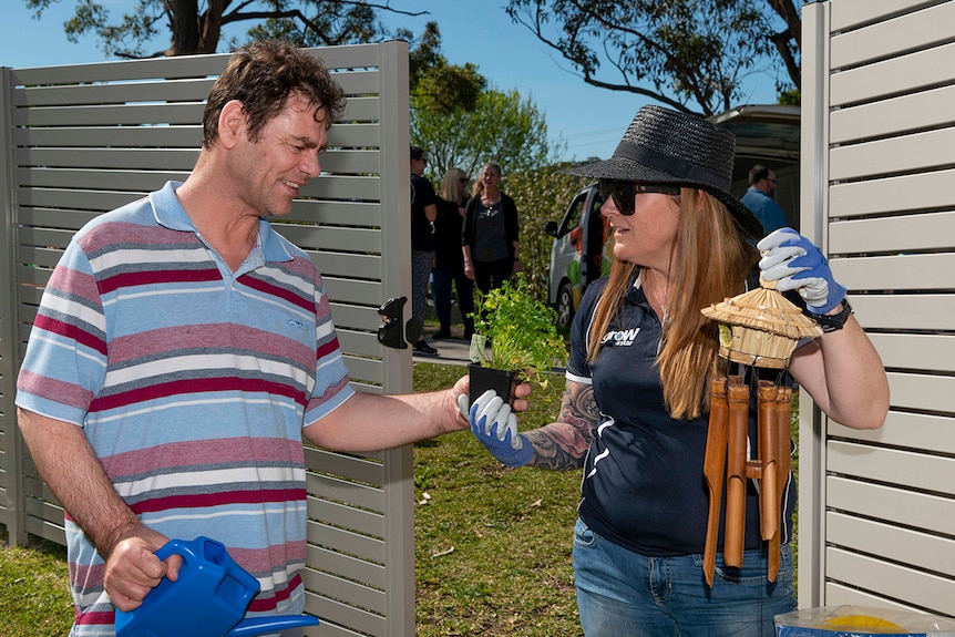 A man and a woman outdoors in a garden holding herbs and a wind chime.