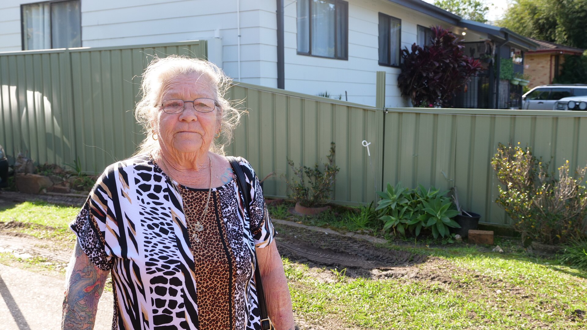 A woman with animal-print clothing and arm tattoos stands in a property with a fence in the background.