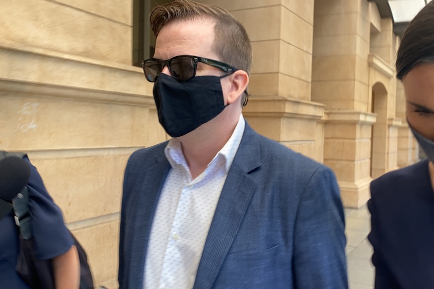 A man wearing sunglasses and a facemask walks outside of a court building
