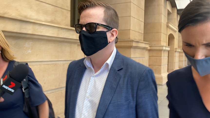 A man wearing sunglasses and a facemask walks outside of a court building