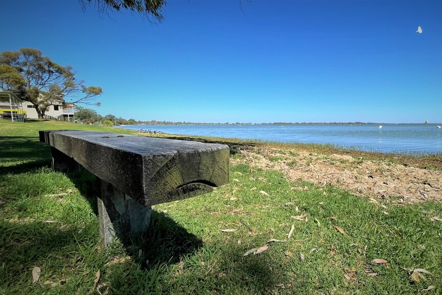 A wooden bench in the foreground, on green grass, with a blue lake and blue skies in the background.