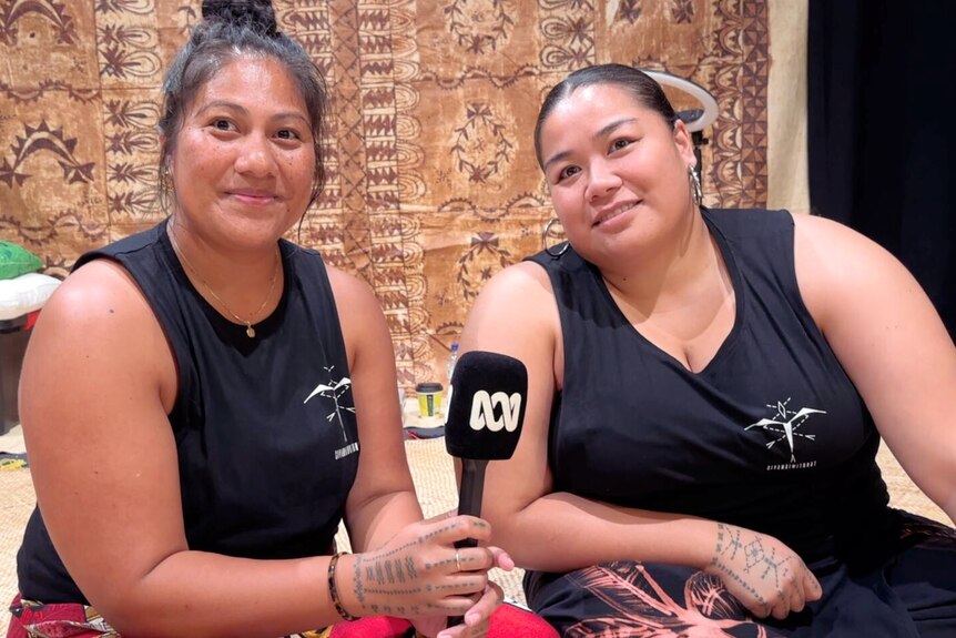 Two Samoan women wear black sleeveless tees and tualima inked on their hands. 