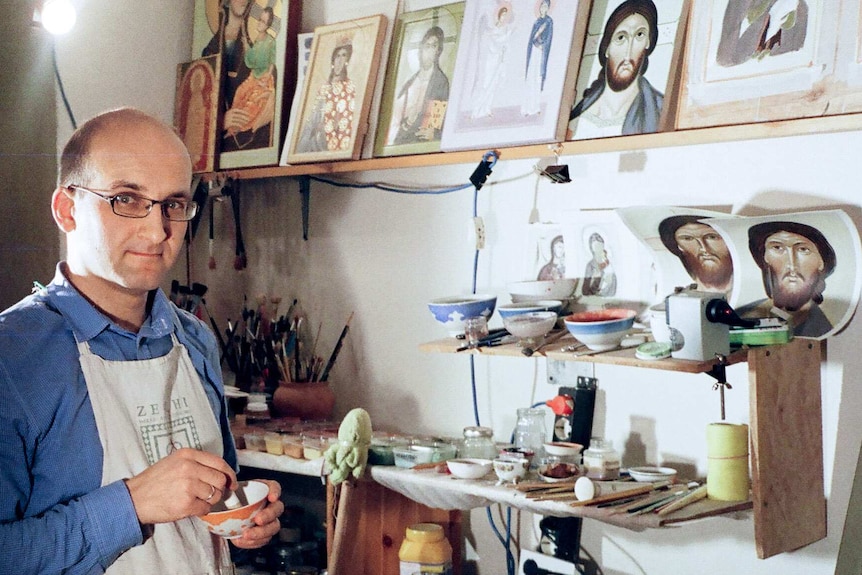 A man in an apron holds his painting equipment while standing in front of images of icons in his studio.