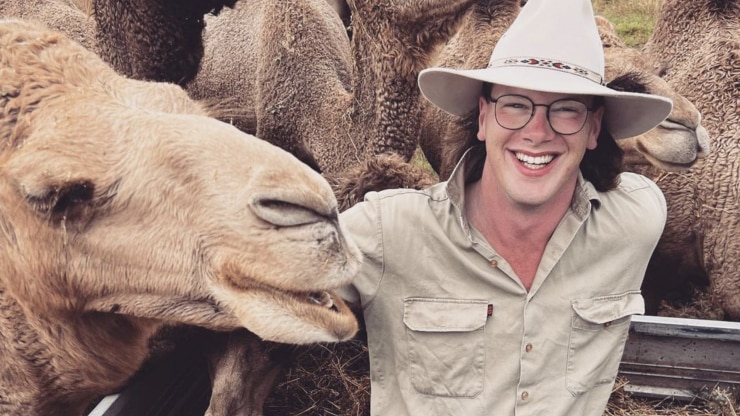 man with long hair and cowboy hat smiling surrounded by camels