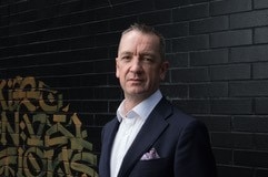 A man, slightly frowning,  in a black suit standing in front of a black brick wall.