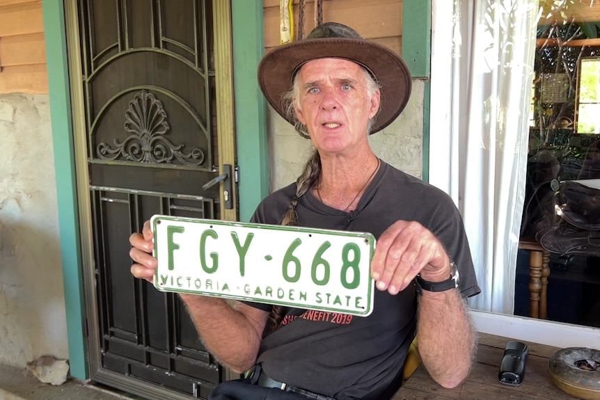 A man with a hat holds up a Victorian number plate FGY668 outside a house