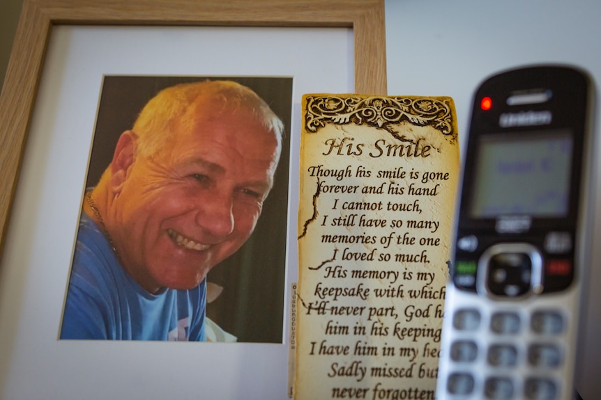 A framed photo of Denise's late husband Ray who is smiling, next to a poem called 'His Smile' and a landline phone.