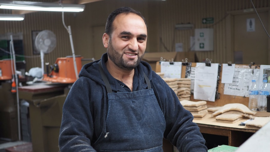 Anas stands at a work bench in a factory with a piece of sheepskin on the bench in front of him.