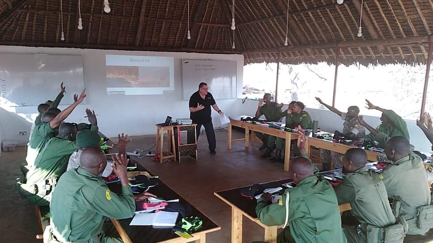A group of rangers in green uniforms sit at desks in a u-shape and hold up their hands while a man stands at the front.
