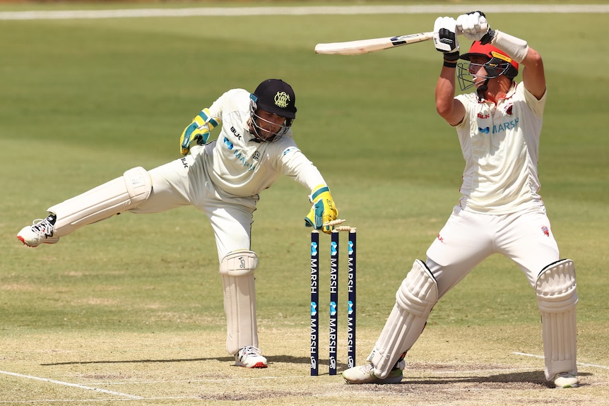 A wicketkeeper stands on one leg as he leans in to whip off the bails while a batsman stands with his bat in the air.