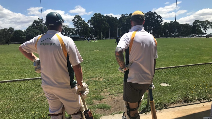 Two cricketers ready to bat are standing at the boundary of the cricket oval in Kinglake, Vicoria. A game of cricket is underway
