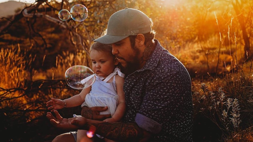 A father sits with a young child, while blowing bubbles.