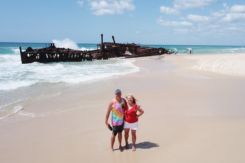 A couple stand on a beach in front of a rusted shipwreck.