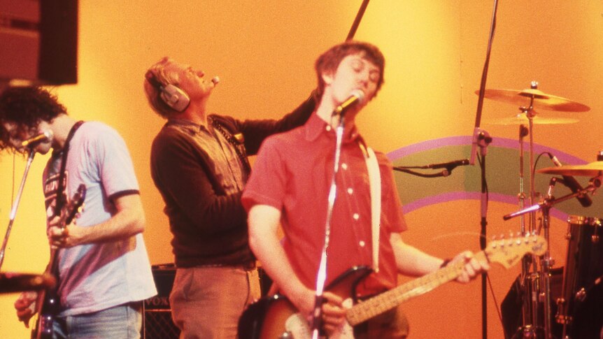Two members of the rock band Custard playing guitars live on a TV set with red and yellow brackgrounds. 