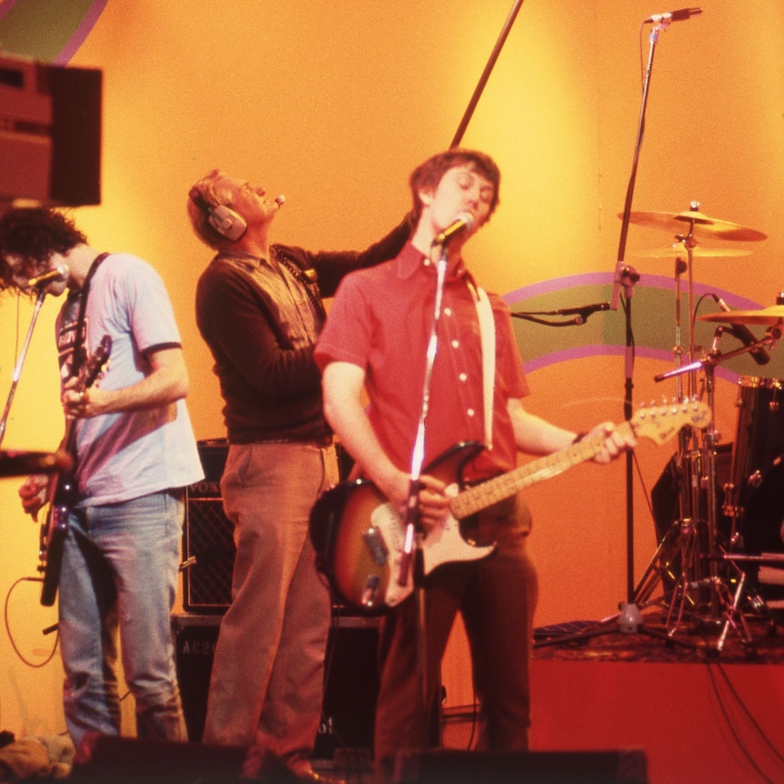 Two members of the rock band Custard playing guitars live on a TV set with red and yellow brackgrounds. 
