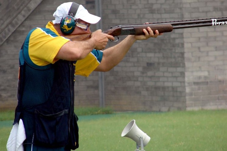 A man in green, gold and black points a gun. He is wearing headphones.