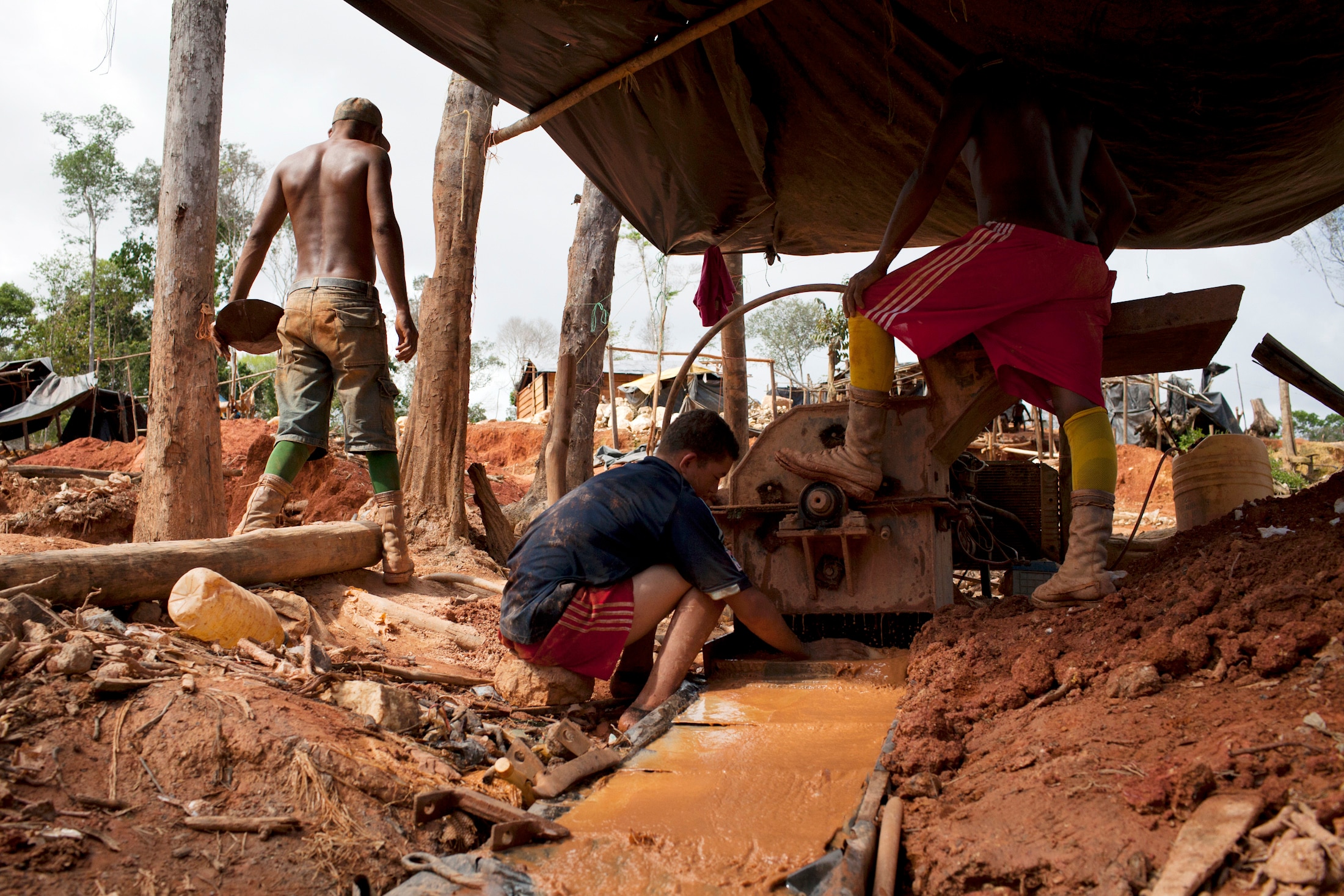 A Venezuean man crouching by a pool of copper orange water in between dirt mounds, in a makeshift shack.