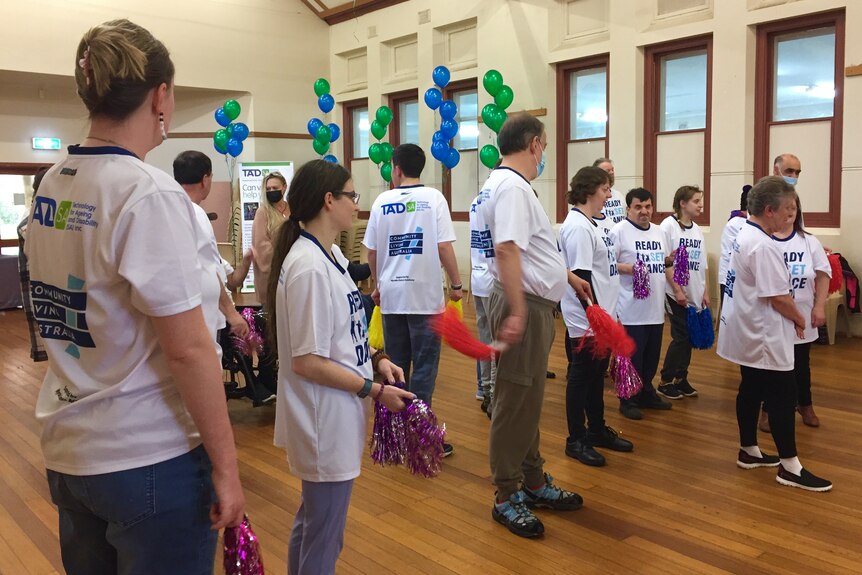 A group of people stand in a hall, holding party streamers, with balloons behind them.