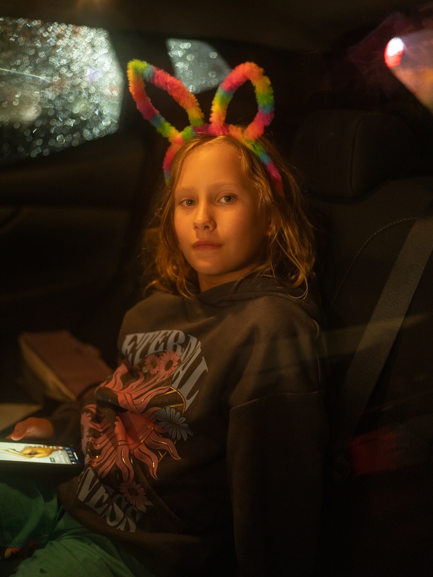 A young girl sitting in the back of a car at night, with rainbow bunny ears on looks to the side with a neutral expression.