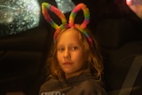 A young girl sitting in the back of a car at night, with rainbow bunny ears on looks to the side with a neutral expression.