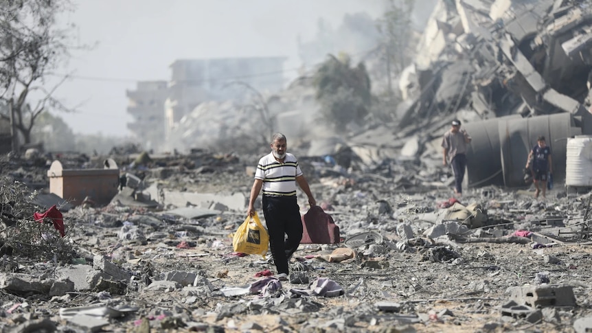 A man carrying a plastic shopping bag walks across rubble and destroyed buildings