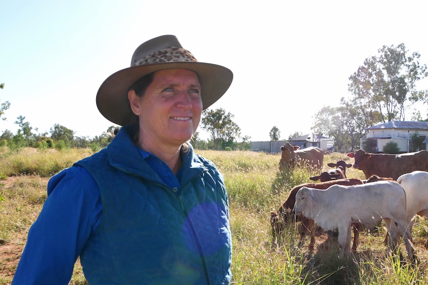 A woman wearing a work shirt and hat smiles. There is cattle in the background
