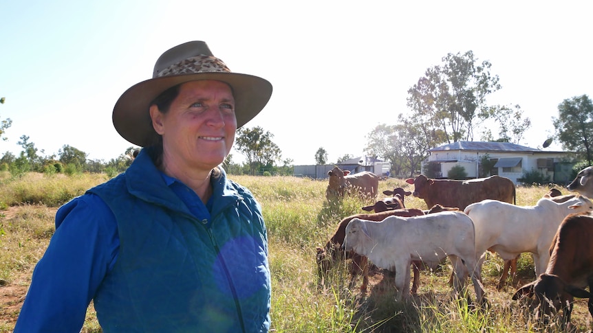 A woman wearing a work shirt and hat smiles. There is cattle in the background