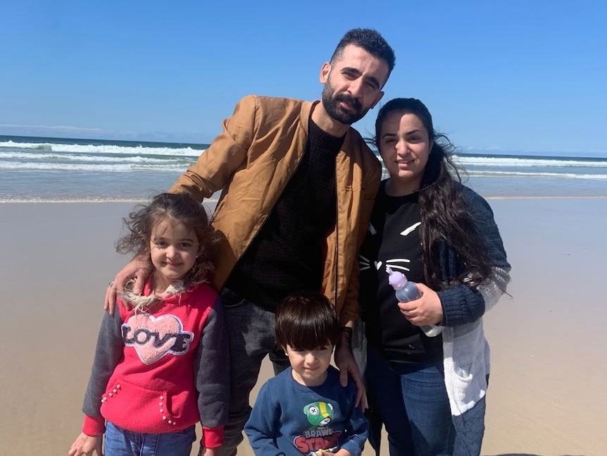 A mother and father with their two children stand with their arms around each other at the beach.