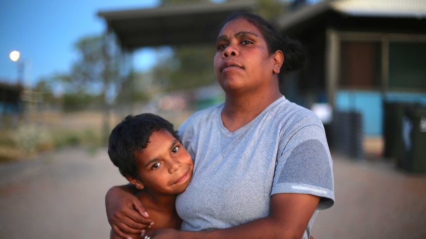 A young Aboriginal boy smiles while he is being hugged my his mother who looks off into the distance.