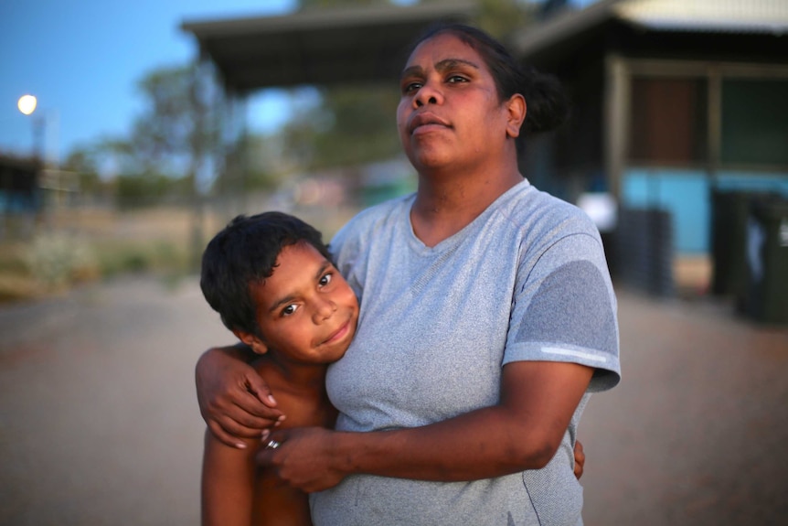 A young Aboriginal boy smiles while he is being hugged my his mother who looks off into the distance.