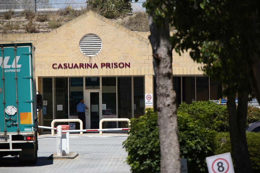 A wide shot of the front entrance to Casuarina Prison, with a truck parked on the left and trees on the right.