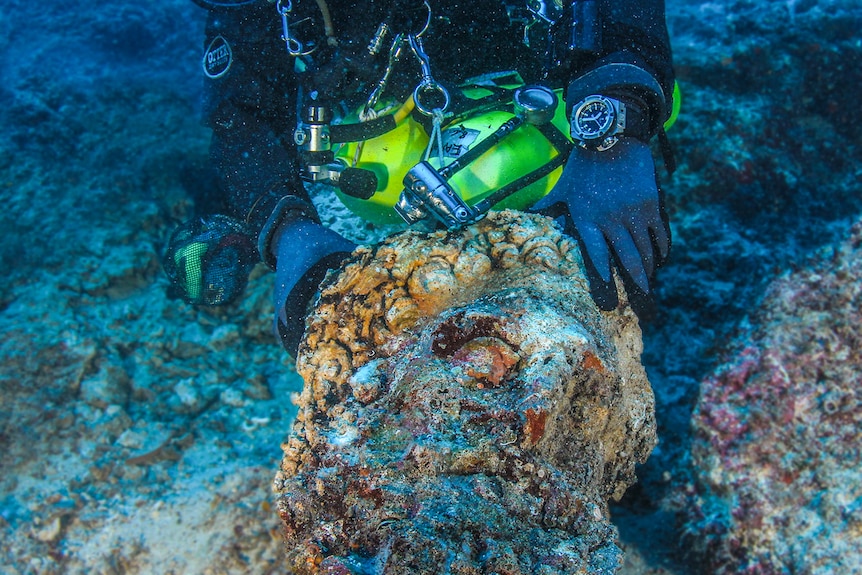 A man wearing a diving suit holds up a marble head covered in grime.