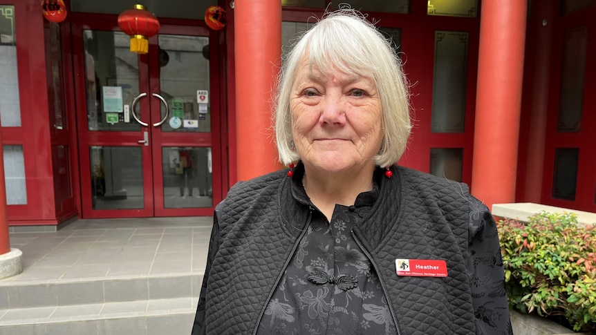 A woman with white jaw length bob wearing a black jacket with a red name tag with Heather in front of red Chinese doors.