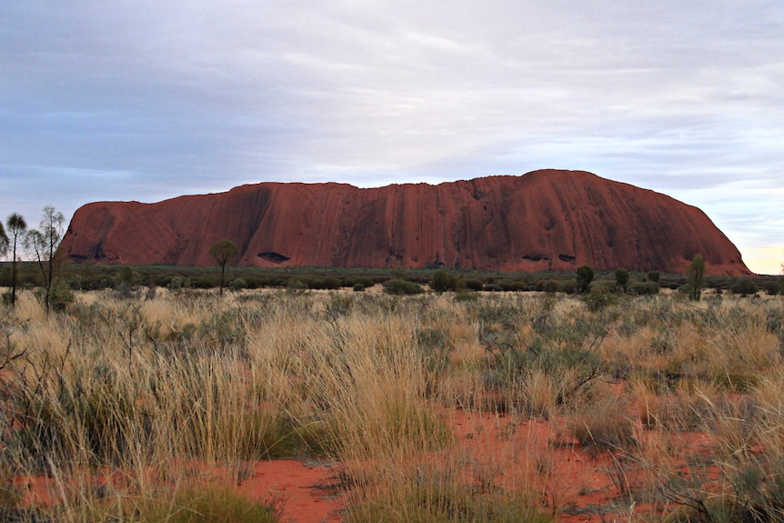 Uluru with cloudy skies in the background and red dirt in the foreground.