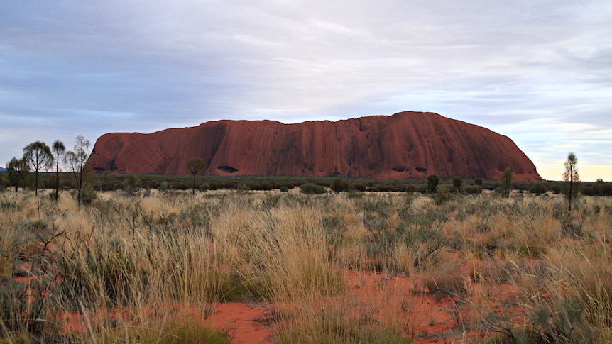 Uluru with cloudy skies in the background and red dirt in the foreground.