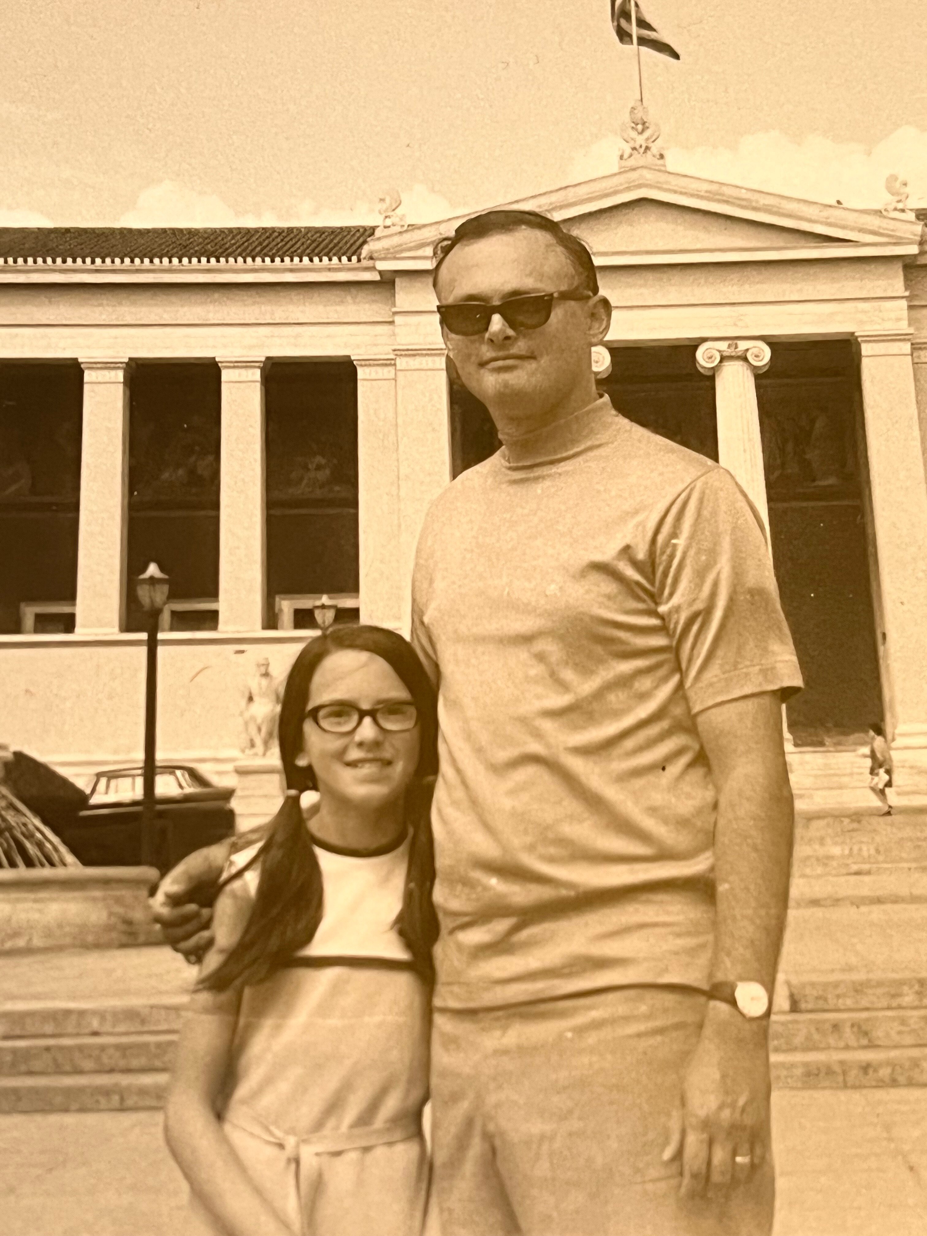 Black and white photo of a young Cynthia Dearborn with plaits standing next to her father, wearing a shirt and sunglasses.