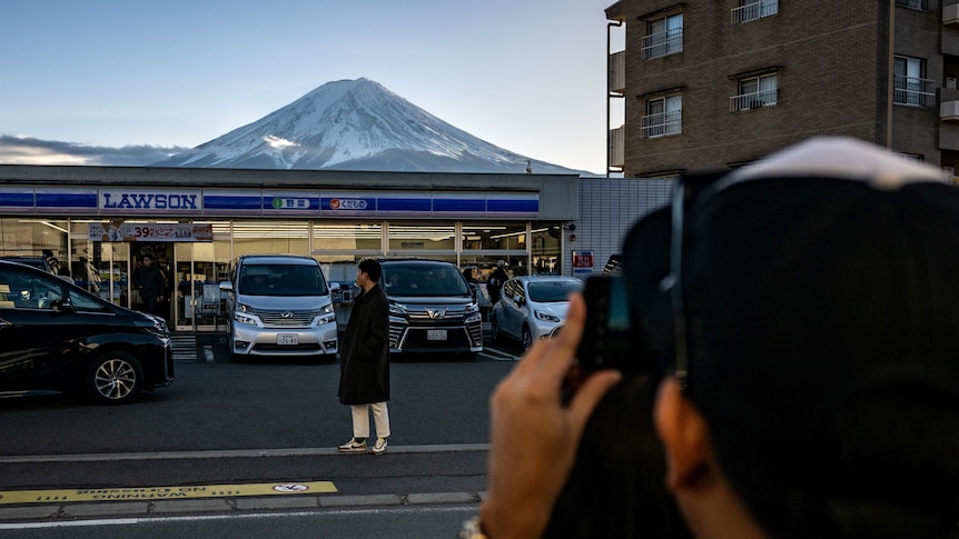 A man poses in front of a service station with Mt Fuji in the background as someone takes a photo